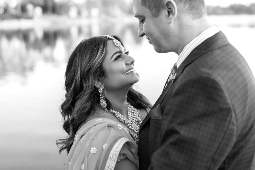 A joyful couple sharing an intimate moment by the water, gazing into each other's eyes with love. The woman, dressed in traditional South Asian attire with intricate jewelry, smiles warmly at her partner, who is wearing a tailored suit. The black-and-white portrait captures their deep connection and timeless romance.